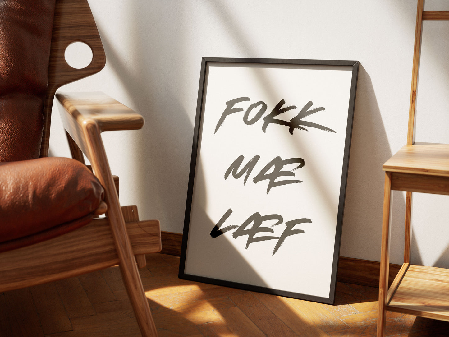 Innrammað plakat upp við hvítan vegg, á milli hillu og viðarstóls. Á plakatinu stendur "Fokk mæ læf"